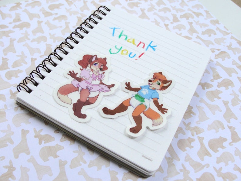 May include: A white notebook with a spiral binding and the words "Thank You!" written in colorful marker. Two cartoon fox stickers are attached to the notebook. One fox is wearing a pink dress and the other is wearing a blue shirt and a diaper.