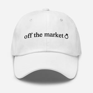 Puede incluir: Gorra de béisbol blanca con bordado negro que dice "off the market" con un símbolo de anillo.