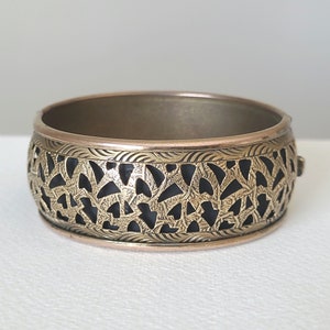 May include: A gold bangle bracelet with a black filigree design. The bracelet is made of metal and has a smooth, polished finish.