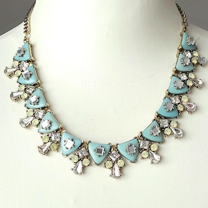 May include: A gold-tone statement necklace featuring a series of light blue triangular resin pieces accented with clear square crystals and small pale yellow round stones, all arranged in a tiered design on a gold-tone metal chain link base.
