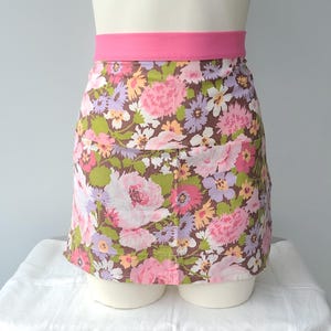 A vinrage floral print apron with a hot pink waistband. The apron features a brown background with a pattern of pink, purple, yellow, and green flowers. The apron is displayed on a mannequin.
