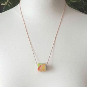 May include: A rose gold chain necklace with a colorful, abstract, rectangular pendant.