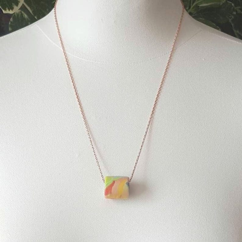 May include: A rose gold chain necklace with a colorful, abstract, rectangular pendant.