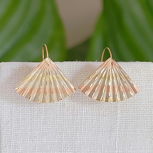 May include: A pair of gold-toned fan-shaped earrings. The earrings have a layered design with alternating stripes of gold, rose gold, and a lighter gold color. The earrings are displayed on a white surface against a blurred green background.