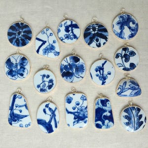 May include: Collection of blue and white porcelain pendants with various floral and nature-inspired designs. Each pendant has a gold-colored metal frame and a loop for hanging. The pendants are in various shapes, including round, teardrop, and rectangular.