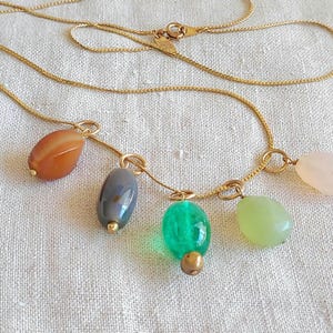 May include: A gold-toned necklace featuring five colorful gemstone pendants. The pendants include orange, gray, green, light green, and pink stones. The necklace is displayed on a white fabric surface.