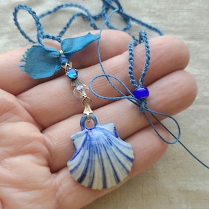 May include: A blue and white porcelain seashell pendant necklace with a blue ribbon and blue beads. The necklace is made with blue cord and has a gold clasp.