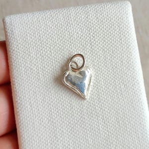 May include: A small, silver heart charm with a loop for attaching to a necklace or bracelet. The heart has a slightly textured surface and a rounded shape. The charm is displayed on a white, textured surface.