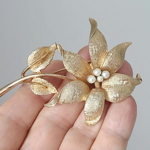 May include: A gold-toned brooch in the shape of a flower with three faux pearl accents in the center. The flower has five petals and is attached to a stem with leaves.