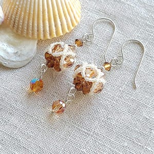 May include: A pair of handcrafted earrings featuring intricate beadwork in shades of brown and cream. The earrings have a floral design with dangling amber-colored crystal beads and silver-tone ear wires. The earrings are displayed on a textured, light-colored surface.