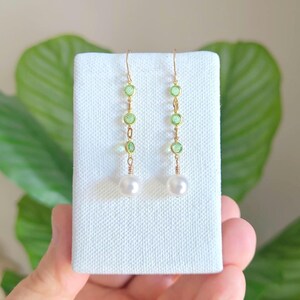 May include: Gold-toned dangle earrings featuring a series of light green gemstones and a single white pearl at the bottom. The earrings are displayed on a white surface against a backdrop of green leaves.