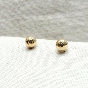 May include: A pair of small, gold-colored stud earrings. Each earring is a textured sphere, reflecting light. The earrings are displayed on a white, textured surface, creating a simple and elegant presentation. These earrings are suitable for everyday wear.