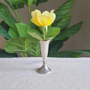 May include: A yellow flower with green leaves in a small, silver-colored vase. The vase has a flared top and a round base. The background features large green leaves and a neutral-colored wall.