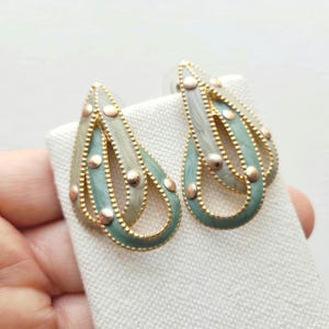 May include: A pair of gold tone earrings with a teardrop design. The earrings are made of two layers of metal, with the top layer being a lighter shade of green and the bottom layer being a darker shade of green. The earrings are decorated with small gold dots.