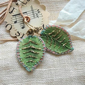 May include: A pair of leaf-shaped earrings crafted from green fabric with gold metallic thread embroidery detailing the veins. The edges are finished with light blue and pink stitching. The earrings feature bronze-toned metal hooks and flower-shaped accents.