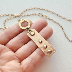 May include: A gold chain necklace with a rectangular pendant. The pendant is made of copper and has a textured surface with three gold dots. The pendant is attached to the chain with a gold ring.