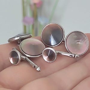 May include: A collection of silver cufflinks with iridescent, pearlescent faces. The cufflinks have a variety of shapes, including round and oval, and are displayed against a soft background. The cufflinks are a classic accessory.