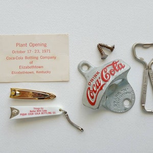 May include: A vintage Coca-Cola bottle opener with a red and white logo. The opener is made of metal and has a silver finish. The text "Drink Coca-Cola" is printed on the opener. There is also a small card with the text "Plant Opening October 17-23, 1971 Coca-Cola Bottling Company of Elizabethtown Elizabethtown, Kentucky".