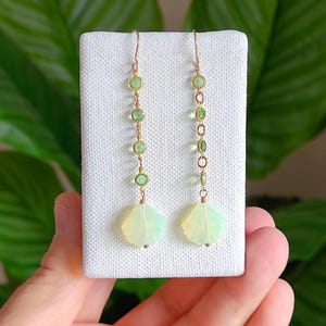 May include: Dangling earrings with a gold-colored wire and chain. The earrings feature a series of small, round, green gemstones and a larger, iridescent, teardrop-shaped stone at the bottom. The earrings are displayed on a white surface.