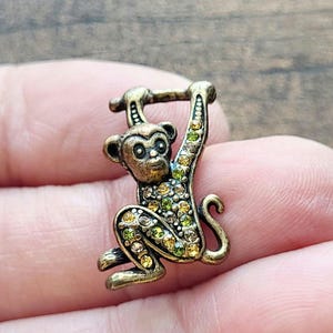 May include: A small, bronze-toned monkey figurine with a detailed design. The monkey is hanging from a bar and is embellished with green and yellow rhinestones. The figurine is held in a person's hand.