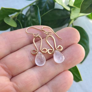 May include: Gold-colored wire earrings with a unique design. Each earring features a curved top, intricate wirework, and a teardrop-shaped, pale pink gemstone. The earrings are displayed against a background of green foliage.