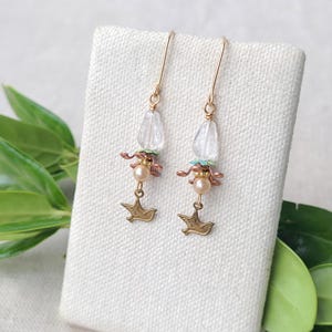 May include: A pair of dangle earrings with gold-colored hooks. Each earring features a clear, faceted bead, pink and green accents, a pearl, and a gold-colored bird charm. The earrings are displayed on a white surface with green leaves.