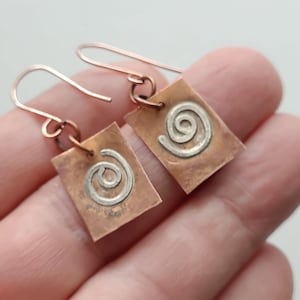 May include: A pair of copper earrings with a silver spiral design. The earrings are square shaped and have a hammered texture.