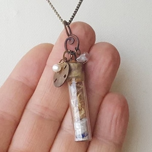 May include: A glass vial necklace with a cork top, filled with dried botanicals and topped with a small brass charm, a pearl, and a pink stone. The necklace is on a brown chain.