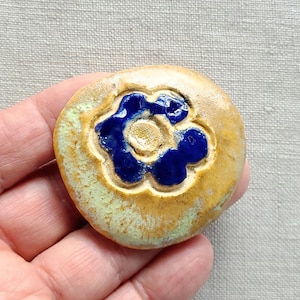 May include: A round, ceramic button with a raised blue flower design in the center. The button has a tan base with a light green edge. The flower has a textured, glossy finish.