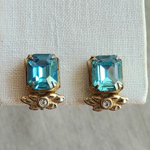 May include: A pair of gold-tone earrings featuring a large, rectangular, emerald-cut light blue stone set above a small, decorative gold-tone leaf motif accented with a single, tiny, clear round rhinestone at the base of each earring.