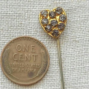 May include: A gold-coloured heart-shaped pin set with small, grey gemstones. A United States one-cent coin is next to the pin, providing a size reference. The background is a textured, light-coloured fabric, highlighting the pin's details.