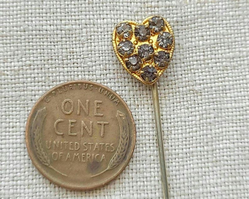 May include: A gold-coloured heart-shaped pin set with small, grey gemstones. A United States one-cent coin is next to the pin, providing a size reference. The background is a textured, light-coloured fabric, highlighting the pin's details.