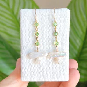 May include: Gold-tone earrings with green gemstones and white baroque pearls. The earrings have a long, dangling design with three green gemstones linked by small gold chains. The bottom of each earring features a large, irregular-shaped white pearl.