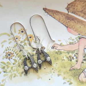 May include: A pair of silver-toned earrings with a unique design. Each earring features a curved, wing-like shape with small, clear crystals and a larger crystal at the center. The earrings have a hook closure. The background is a vintage-style illustration.