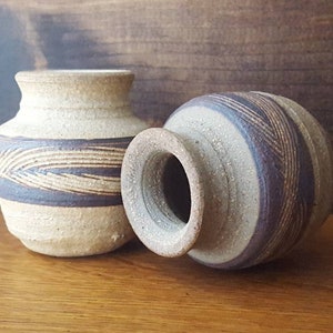 May include: Two small, brown and white ceramic vases with a striped design. The vases are sitting on a wooden surface.