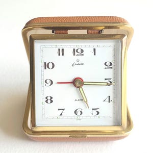 May include: A vintage Endura travel alarm clock with a gold-tone metal frame and a light brown leatherette case. The white clock face has black numerals, a red second hand, and gold-tone hour and minute hands. The word "ALARM" is printed on the face.