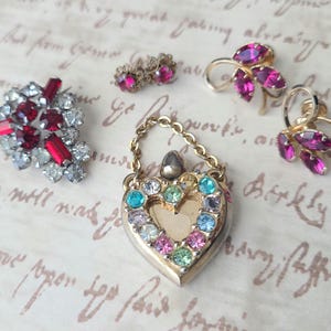 May include: A set of four vintage jewelry pieces featuring red and pink rhinestones. The set includes a heart-shaped locket with a chain, a pair of earrings with a leaf design, and a brooch with a cluster of red rhinestones.