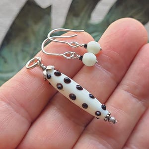 May include: A pair of handmade earrings and a pendant. The earrings have silver hooks with small white beads and black accents. The pendant is a white, cylindrical bead with dark brown polka dots and silver accents.