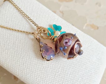 May include: A gold-toned necklace with three charms. One charm is a teardrop-shaped pendant with a purple stone. Another is a round charm with a dog-like face, and the third is a turquoise flower with a pearl center.