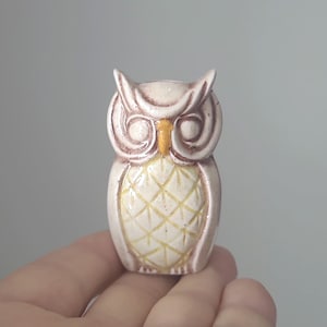 May include: A small ceramic owl figurine with brown and white paint. The owl has yellow eyes and a detailed pattern on its chest.