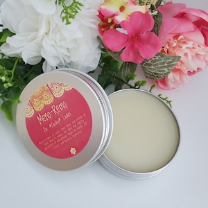 May include: An open, silver-colored tin of Meno-Remo balm for flashy ladies, with a red and gold label. The balm is light yellow. The tin sits next to a bouquet of pink and white flowers with green foliage.