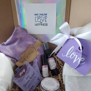 May include: A gift box filled with self-care items, including a lavender eye pillow, a bottle of essential oil, a jar of balm, and two tea bags. The box is decorated with a white ribbon and a card that says "Sending you love".