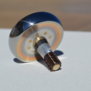 May include: A silver spinning top with a gold and copper ring around the edge. The top has a silver and copper stem with a gold tip.