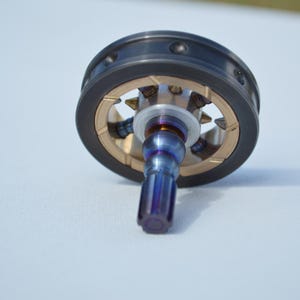 May include: A metal object with a dark grey outer ring and a gold-coloured inner ring. The object has a blue and purple stem. The object is on a light blue surface.