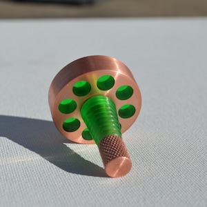 Może przedstawiać: Drewniany bączek z miedzianym wykończeniem, z zielonym środkiem i szeregiem otworów wokół krawędzi.