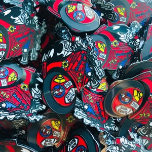 Puede incluir: Primer plano de un montón de pines de esmalte rojo, negro y blanco. Los pines representan a un personaje de dibujos animados conduciendo un coche rojo con el texto "Bada Bing!" en el lateral. Los pines son de diferentes tamaños y formas.