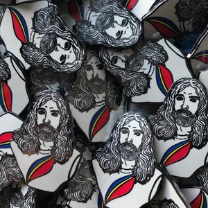 Puede incluir: Un conjunto de pines esmaltados con una ilustración estilizada de un hombre con pelo largo y barba. Los pines tienen un fondo blanco con un diseño rojo, amarillo y azul en el pecho. Los pines son en blanco y negro.