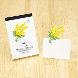 May include: A white Diecut Stand Message Pad and a white message card with a yellow floral design. The pad has a black top and the text "Diecut Stand Message Pad" and "Marumond Made in Japan". The floral design is a bouquet of yellow flowers.