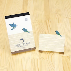 May include: A Diecut Stand Message Pad featuring a blue bird design. The notepad has a black top and a light wood grain pattern. A smaller stand-alone notepad with a matching design is also present.