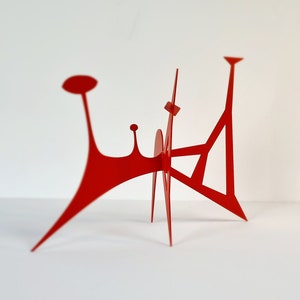 May include: A red metal sculpture with abstract shapes and a geometric design. The sculpture has a tall, thin base and a curved top.